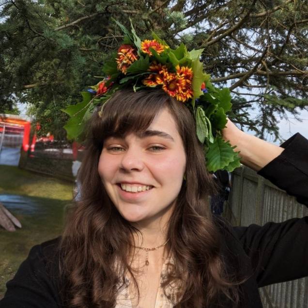 Alina Onysko Specjalista ds. widoczności, młoda uśmiechnięta kobieta na tle iglastego drzewa, lewą ręką przytrzymuje na głowie wianek z kwiatów