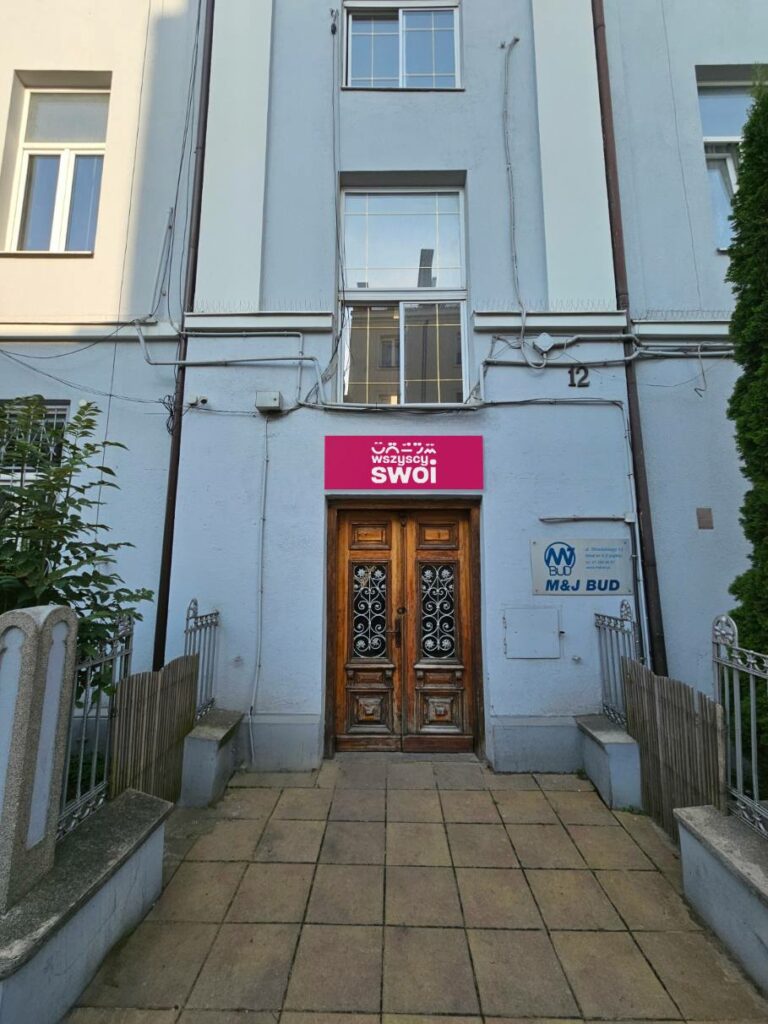 brązowe drzwi do starej kamienicy o szaro-niebieskiej elewacji, nad którymi wisi szyld z białym logo Wszyscy Swoi na fuksjowym tle