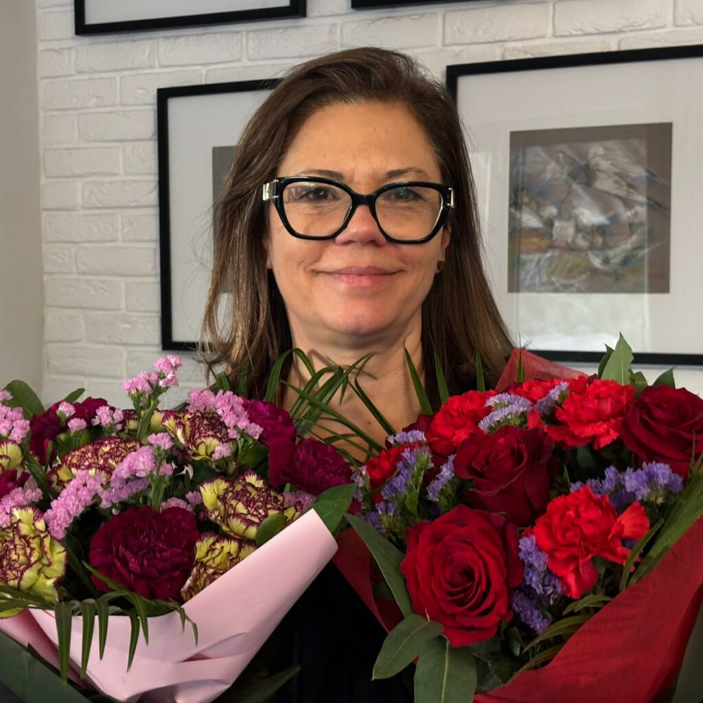 Dorota Slapek - pogodna, młoda kobieta w okularach trzyma przed sobą dwa bukiety kwiatów