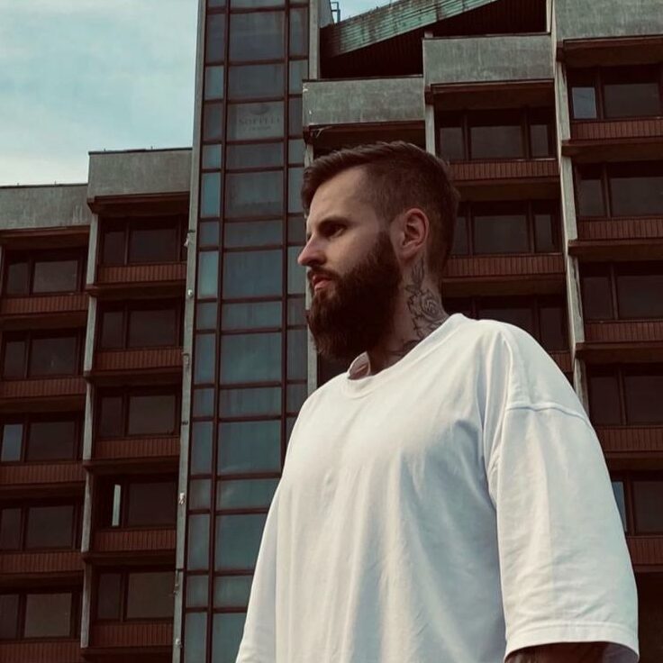 Wojtek Piwoński - młody człowiek z brodą i tatuażem na szyi, ubrany w białą koszulkę stoi na tle modernistycznego budynku mieszkalnego; zdjęcie z boku, z żabiej perspektywy