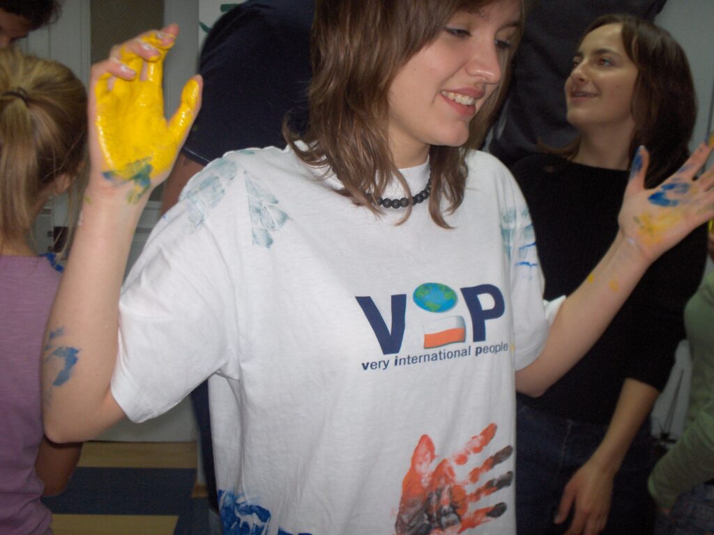 młoda dziewczyna w koszulce z logo VIP w trakcie zabawy grupowej z farbami; ręce w farbie i ślady odbitych rąk innych osób na koszulce