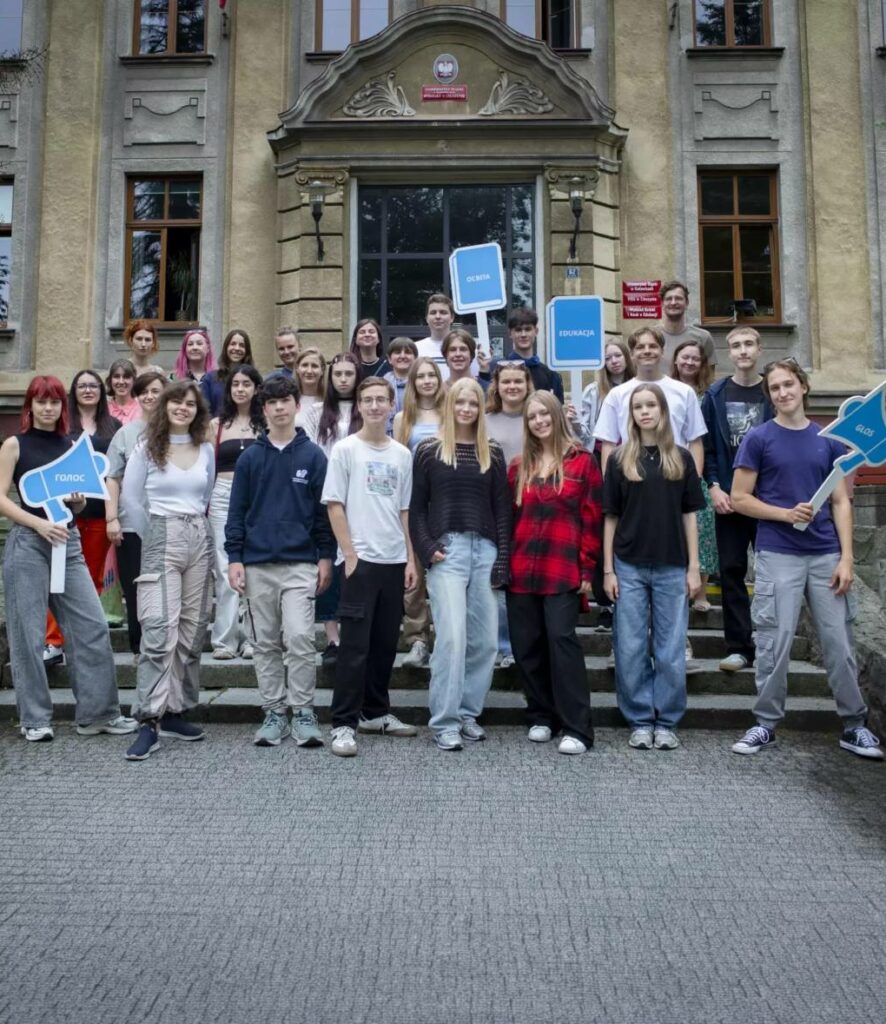 grupa młodzieży z opiekunami stoi na tle starego budynku uniwersytetu z transparentami"edukacja", głos" po polsku i ukraińsku