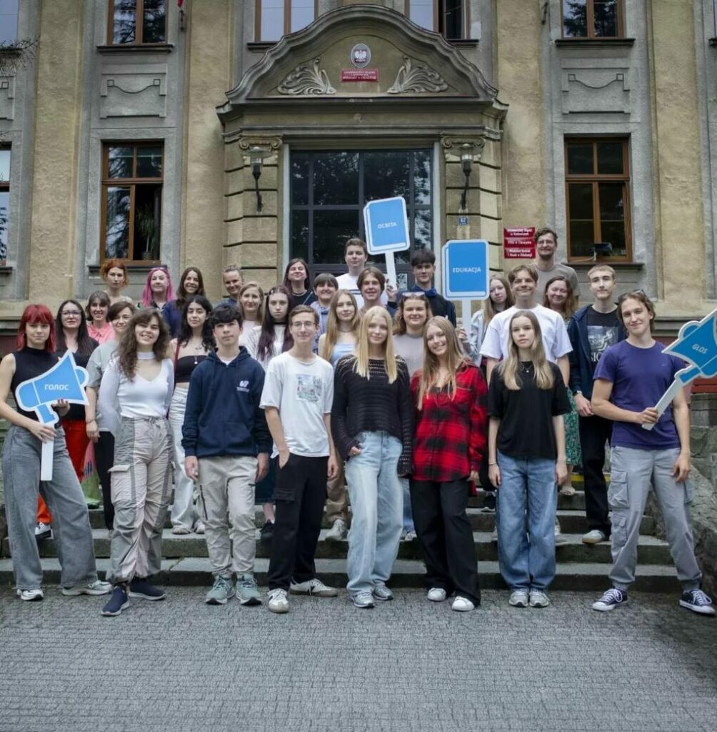 grupa młodzieży z opiekunami stoi na tle starego budynku uniwersytetu z transparentami"edukacja", głos" po polsku i ukraińsku