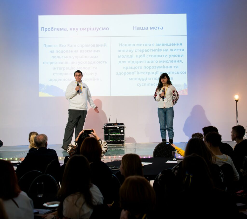 młodzież prowadzi prezentację na scenie; w tle obraz z projektora