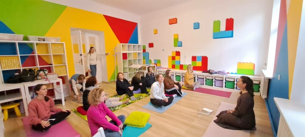 Grupa kobiet siedzących na matach podczas sesji medytacji Kundalini w jasnej sali o kolorowych ścianach ozdobionych motywem klocków Lego.