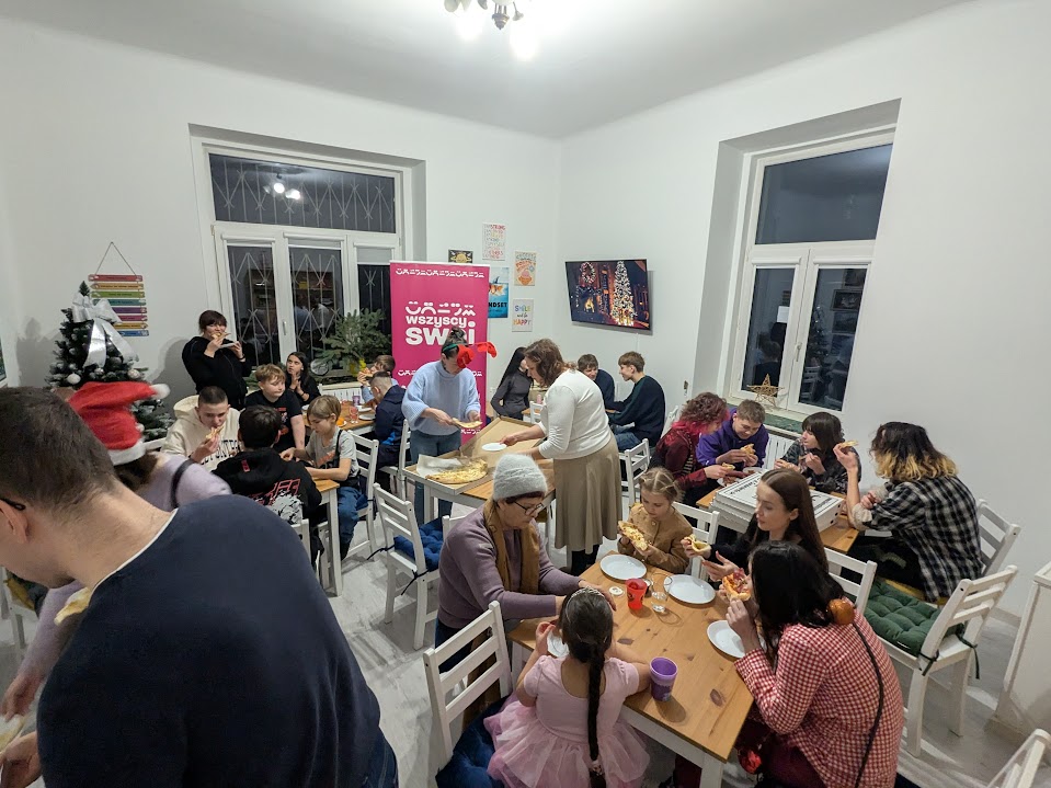Uczestnicy świątecznego spotkania siedzą przy stołach w jasnej sali, jedząc pizzę i rozmawiając; w tle widać choinkę oraz różowy baner Wszyscy Swoi.