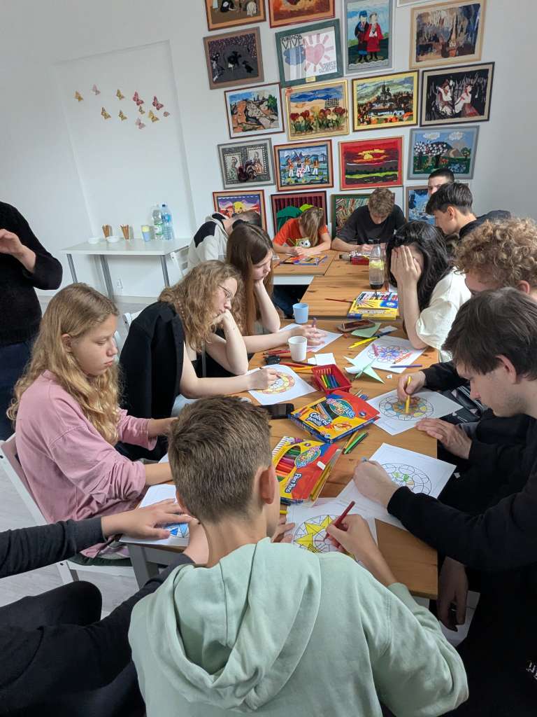 Uczestnicy warsztatów siedzą gęsto przy podłużnym stole i z zaangażowaniem kolorują kredkami oraz pisakami geometryczne mandale na białych kartkach. Na ścianie w tle wisi gęsto rozmieszczona galeria kolorowych, oprawionych obrazów.