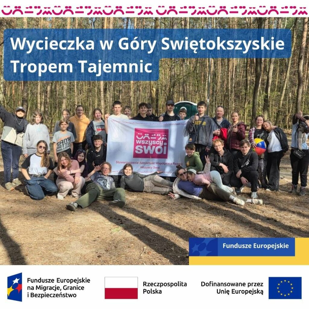 Plakat informacyjny z napisem „Wycieczka w Góry Świętokrzyskie Tropem Tajemnic”. W tle duże zdjęcie grupowe uczestników wycieczki stojących w lesie i trzymających białą flagę z różowym logo i napisem „wszyscy swoi”. Na dole plakatu znajdują się logotypy funduszy unijnych oraz flaga Polski z podpisami instytucji współfinansujących.