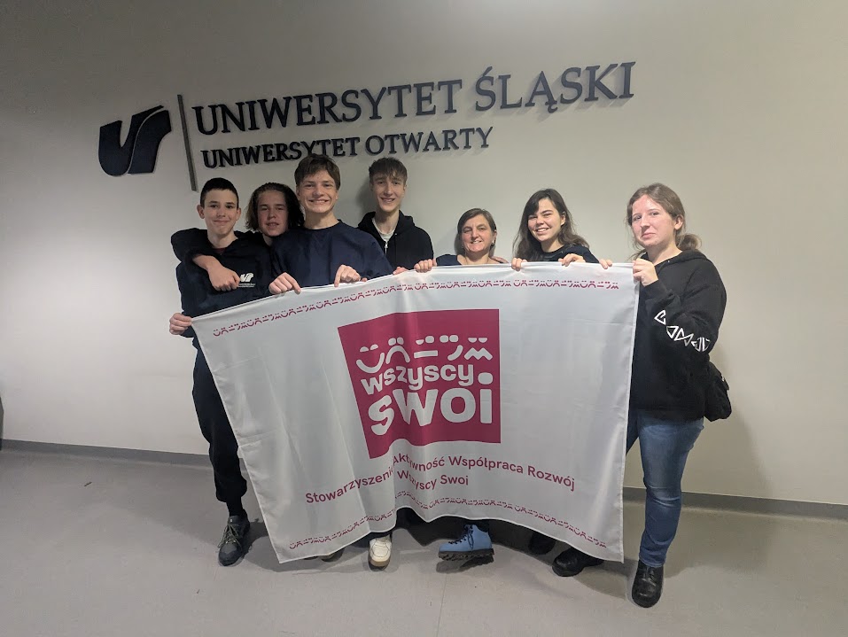 Grupa siedmiu osób pozuje do zdjęcia na tle ściany z napisem Uniwersytet Śląski, Uniwersytet Otwarty. Wspólnie trzymają białą flagę z różowym logo Stowarzyszenia Wszyscy Swoi