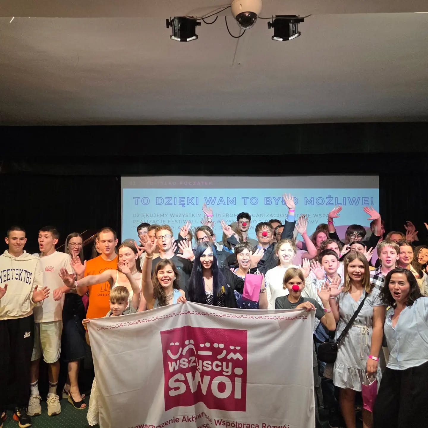 Zdjęcie grupowe uczestników i organizatorów Festiwalu Idei trzymających banner z logo Stowarzyszenia Wszyscy Swoi na scenie Pałacyku Zielińskiego.
