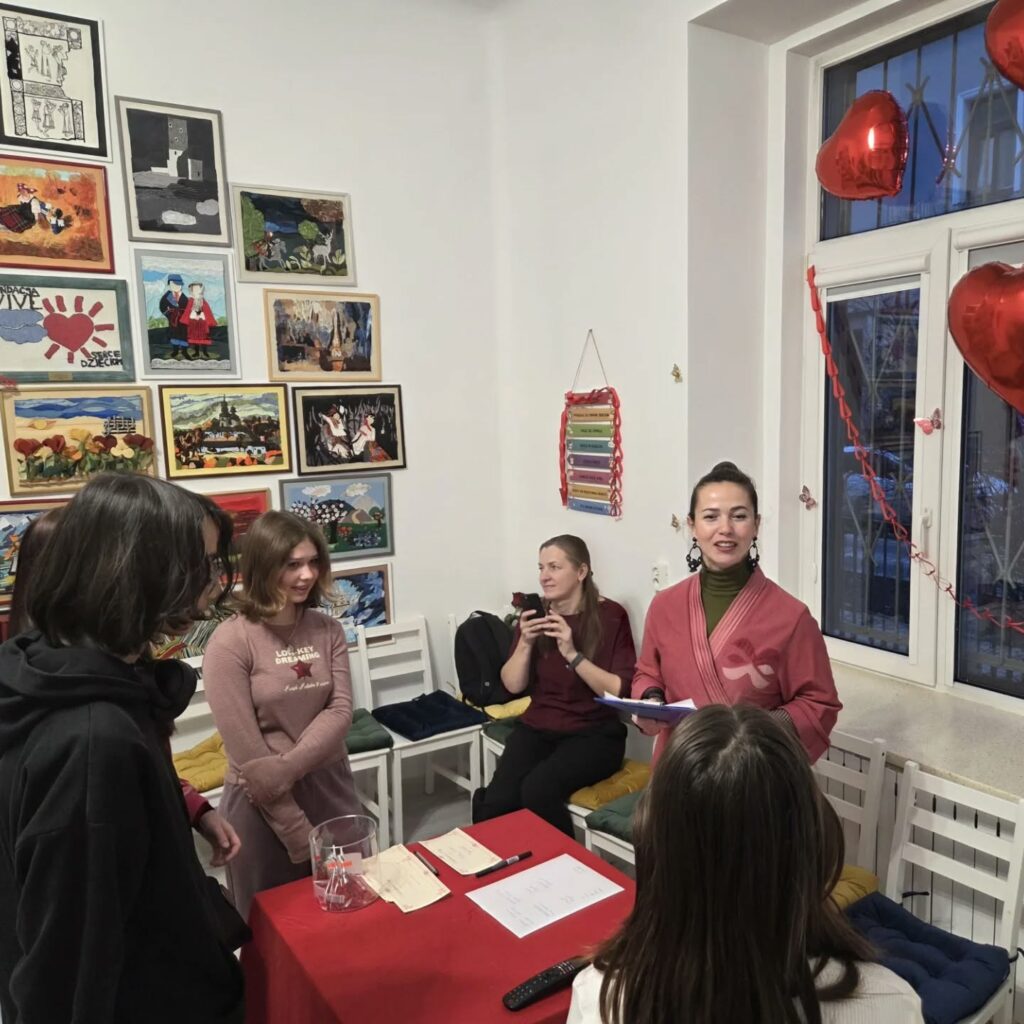 Grupa osób zgromadzona w jasnym pomieszczeniu ozdobionym pracami plastycznymi i czerwonymi balonami w kształcie serc. Po prawej stronie stoi kobieta w różowym kimonie, trzymająca podkładkę z kartką, która przemawia do pozostałych obecnych. W tle siedzi kobieta robiąca zdjęcie smartfonem. Przeznaczone na losy lub małe karteczki naczynie stoi na stole przykrytym czerwonym obrusem.