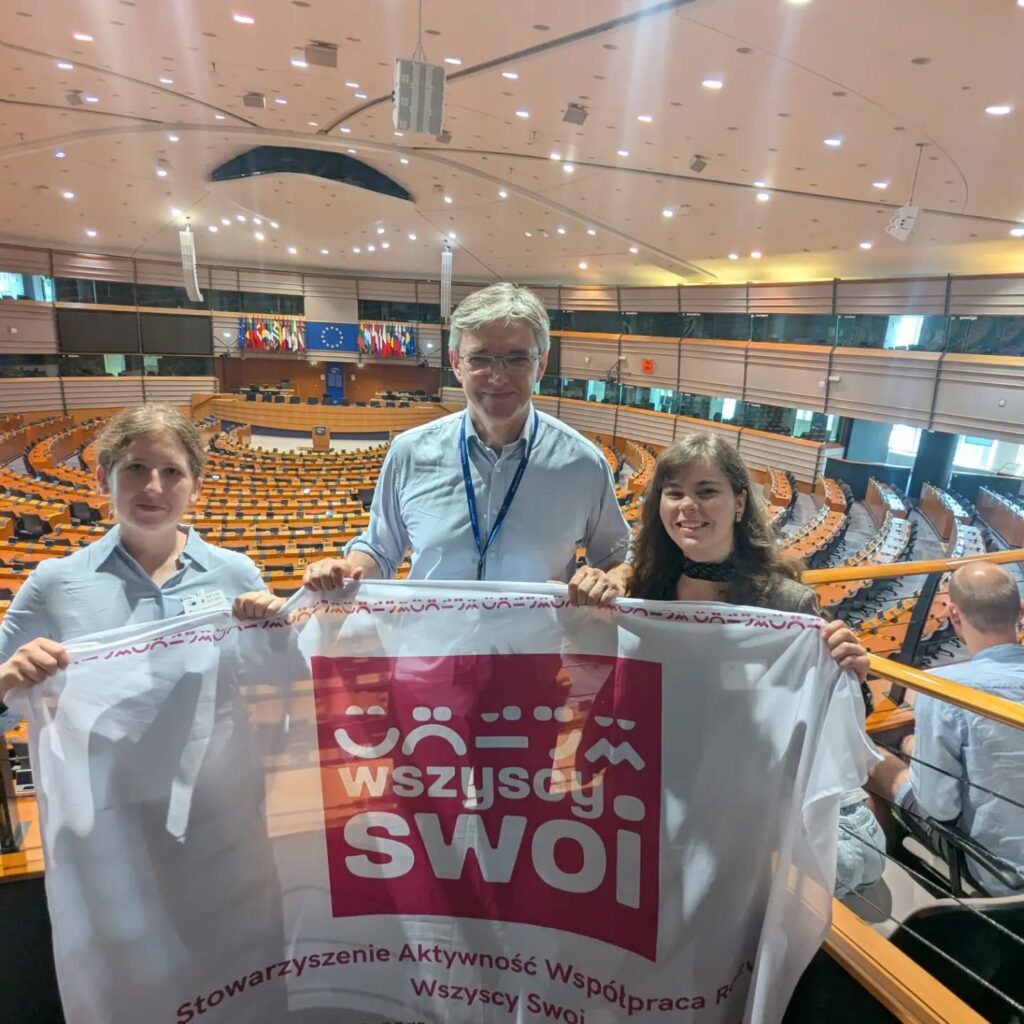 Dwie kobiety wraz z posłem do Parlamentu Europejskiego Adamem Jarubasem trzymający biały baner Stowarzyszenia Wszyscy Swoi na balkonie widokowym wewnątrz sali plenarnej Parlamentu Europejskiego.